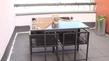 Ein Tisch mit Glasplatte und Metallgestell, begleitet von zwei Stühlen, steht auf einer Terrasse mit gefliestem Boden.
