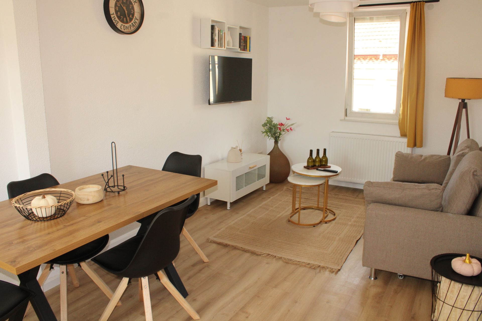 Ein modernes Wohnzimmer mit einem Holzespeisetisch, schwarzen Stühlen, einem weißen Sofa, einem kleinen Beistelltisch, einem an der Wand montierten Fernseher und einem Fenster mit Vorhängen.