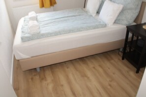 A single bed with a white mattress and a light blue cover, positioned against a wooden floor.