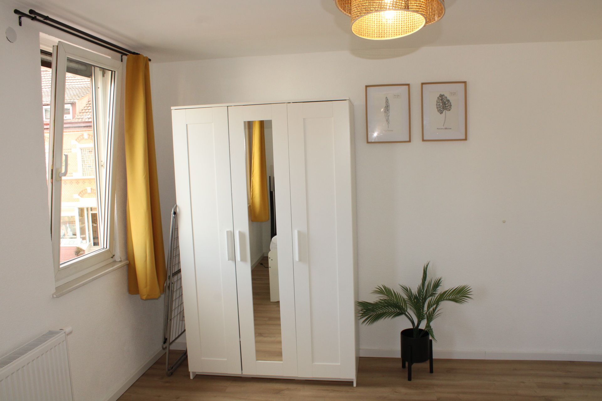 Ein Zimmer mit einem weißen Kleiderschrank, einem Fenster mit gelben Vorhängen, einer Topfpflanze und zwei gerahmten Pflanzenzeichnungen an der Wand.
