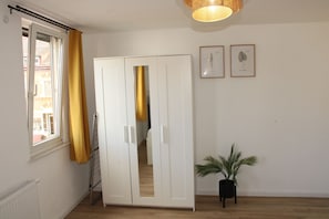 A room with a white wardrobe, a window with yellow curtains, a potted plant, and two framed botanical illustrations on the wall.