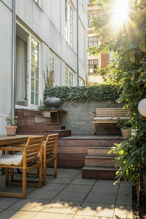 An outdoor patio area with wooden furniture, a stone wall with climbing plants, and a bench. - Two-storey flat with private garden (København S)