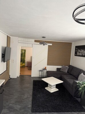 Ein modernes Wohnzimmer mit einem grauen Sofa, einem weißen Couchtisch und einem schwarzen Teppich. An der Wand ist ein Fernseher angebracht, eine Deckenleuchte hängt von der Decke und ein Fenster mit Jalousien ist vorhanden.