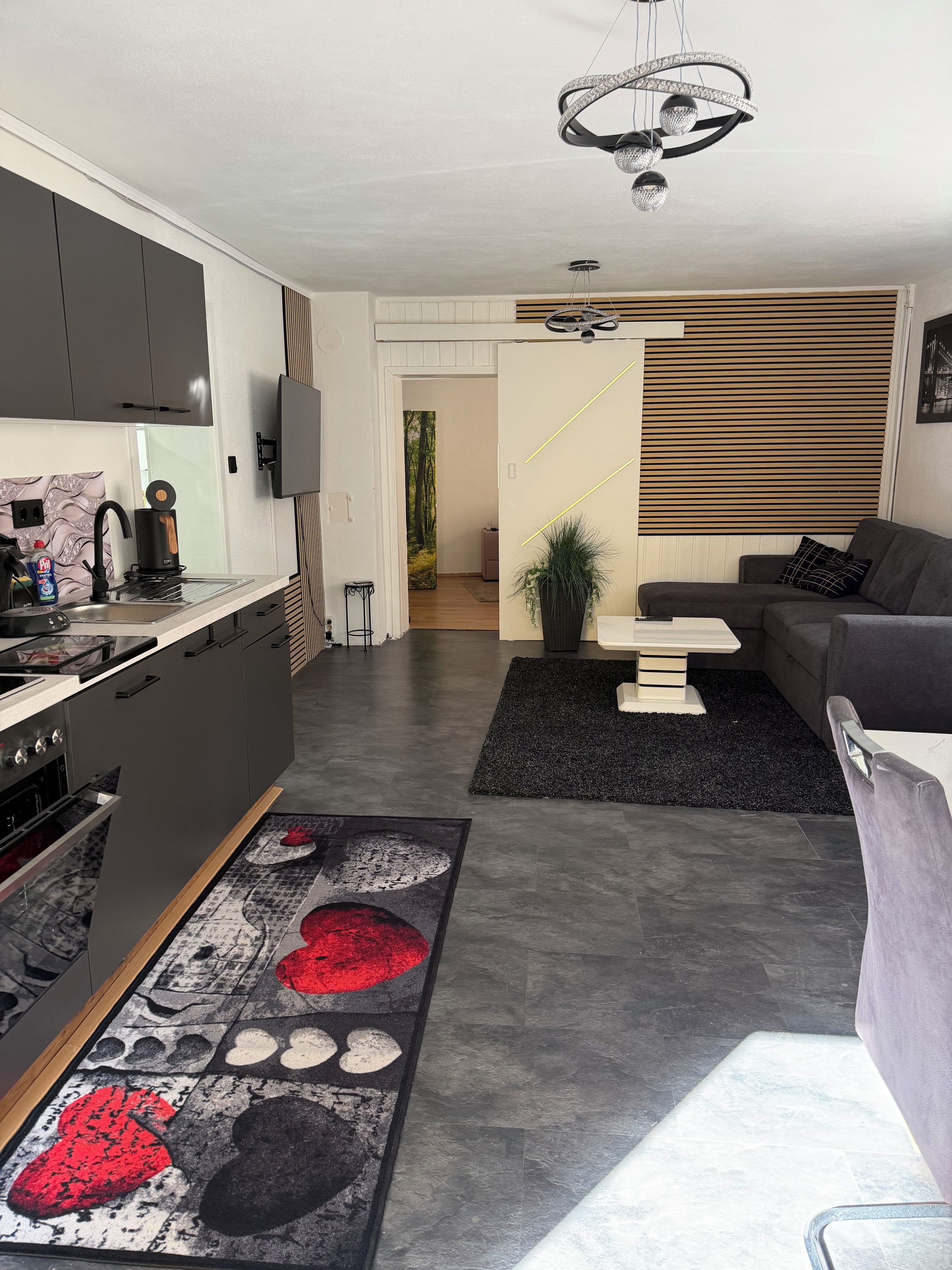 Eine moderne Küche mit Einbauherd, ein Essbereich mit einer Couch und einem Couchtisch sowie ein Wohnzimmer mit großem Fenster.