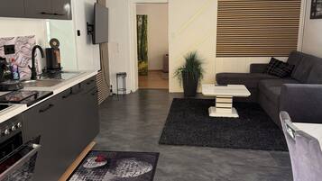 Eine moderne Küche mit Einbauherd, ein Essbereich mit einer Couch und einem Couchtisch sowie ein Wohnzimmer mit großem Fenster.