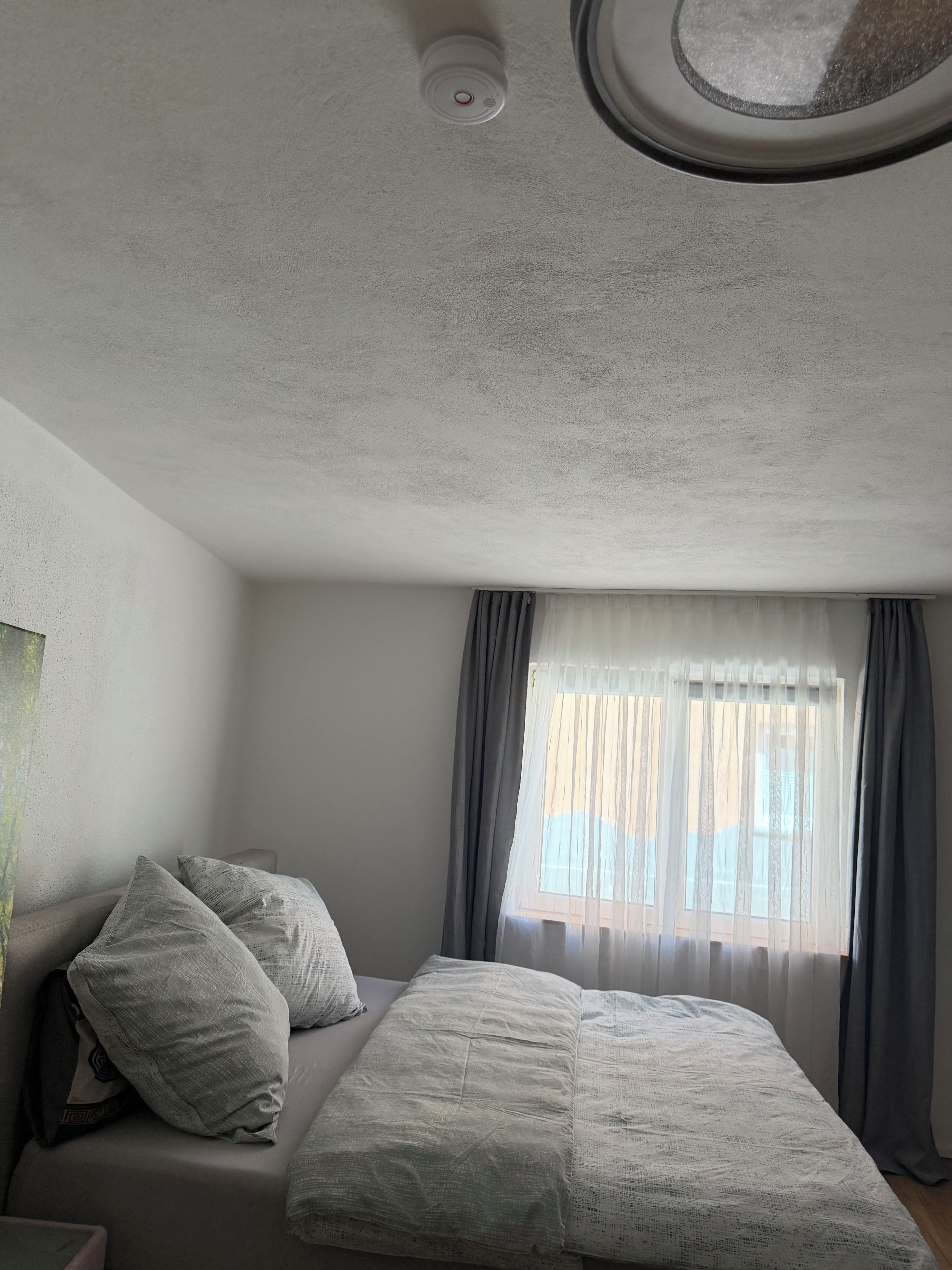 Ein Schlafzimmer mit einem Bett, Kissen, einem Fenster mit Vorhängen und einer Deckenleuchte.