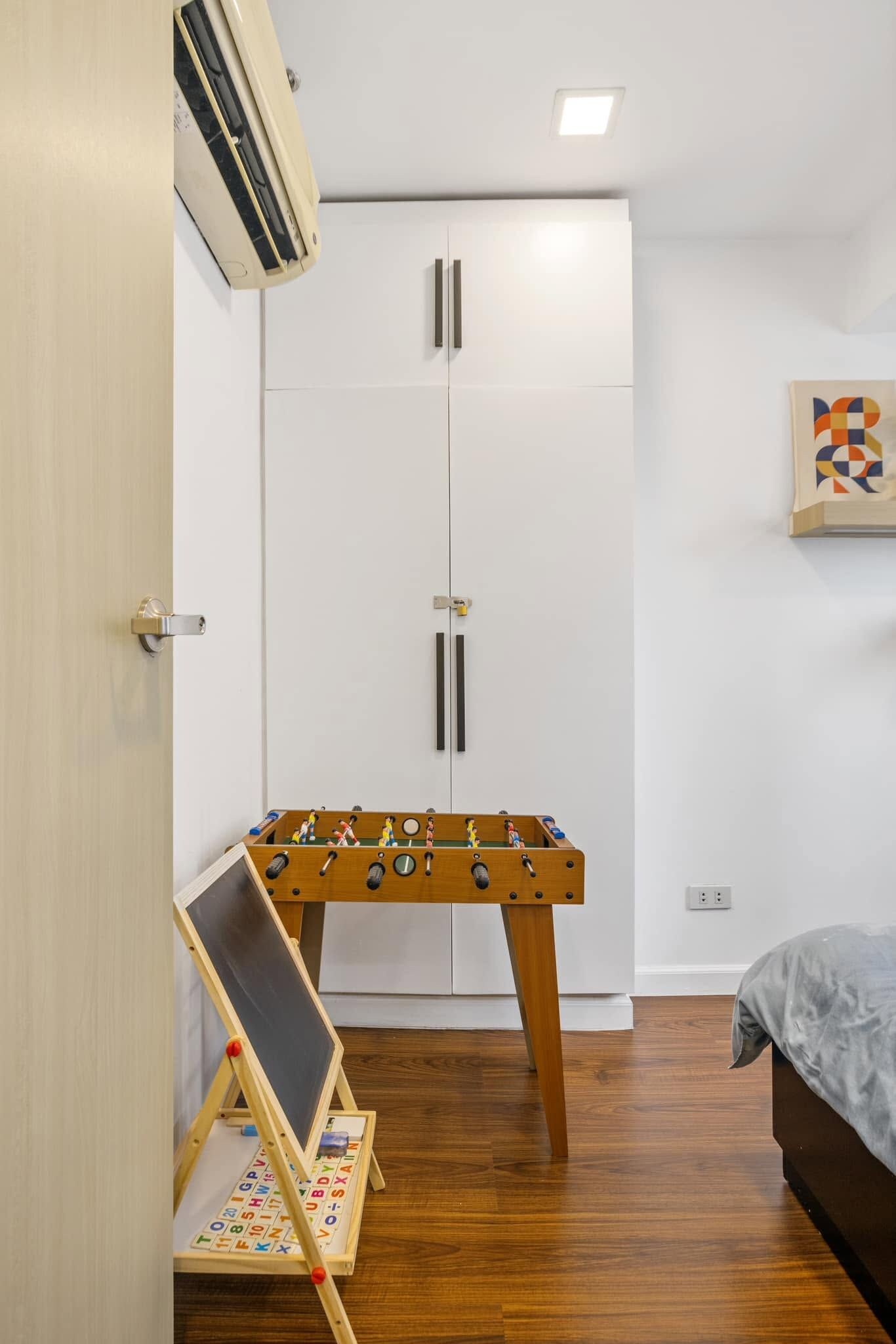 A bedroom with a bed, a wardrobe, a foosball table, a small chalkboard, and a wall-mounted artwork.