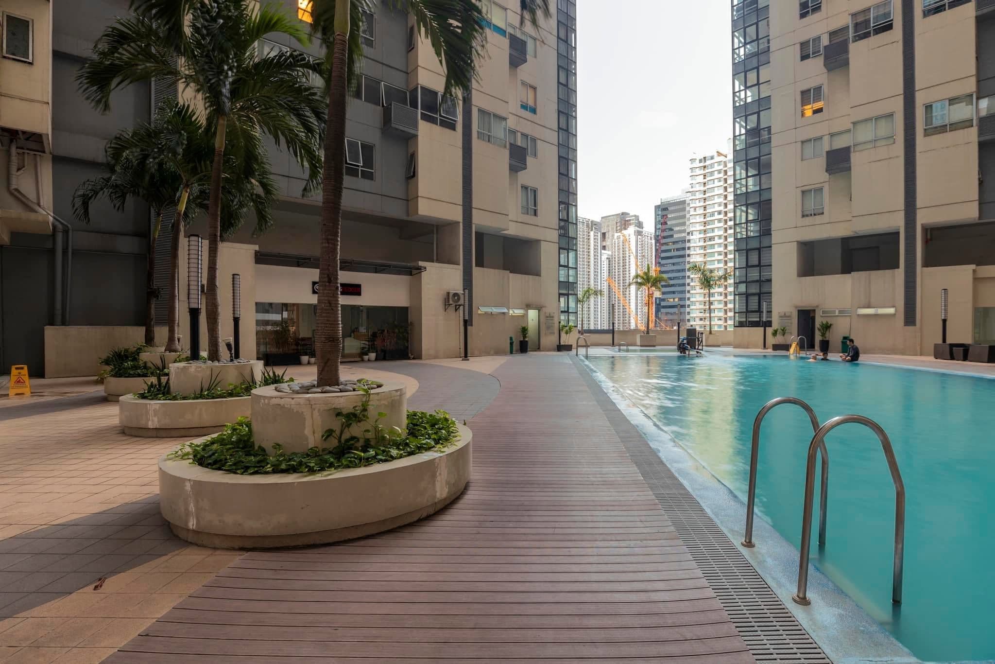 A swimming pool area with a wooden deck, palm trees, and modern apartment buildings in the background.