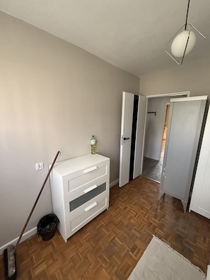 A room with a wooden floor, a white dresser, a mop, a trash bin, and a door leading to another room.