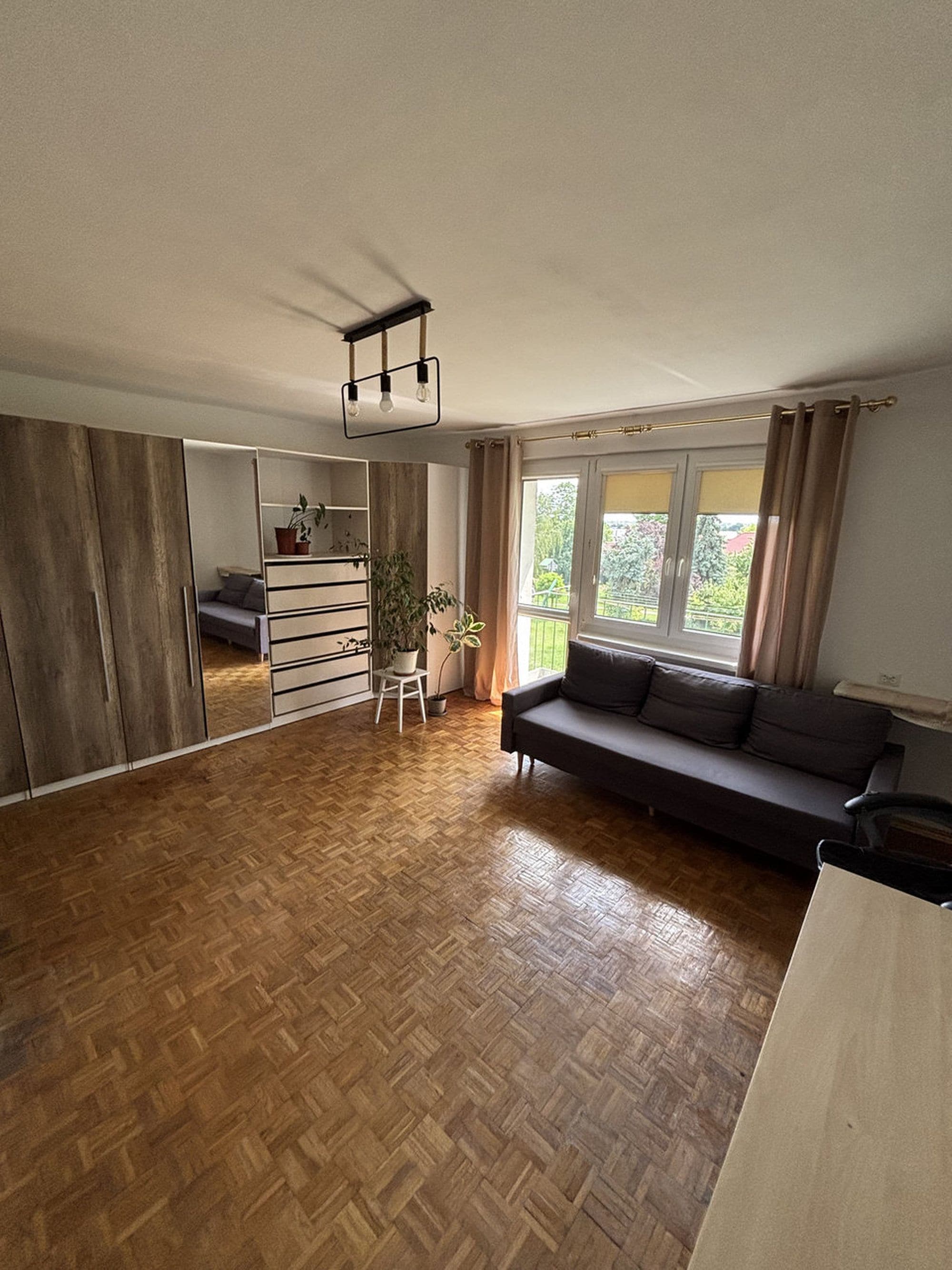 A spacious living area with a wooden parquet floor, a dark-colored sofa, a small white table with a plant, and a wardrobe with sliding doors.