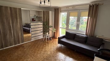 A spacious living area with a wooden parquet floor, a dark-colored sofa, a small white table with a plant, and a wardrobe with sliding doors.