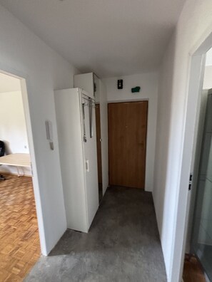 Un pasillo estrecho con una puerta de madera al final, un refrigerador blanco y una pequeña señal verde en la pared.