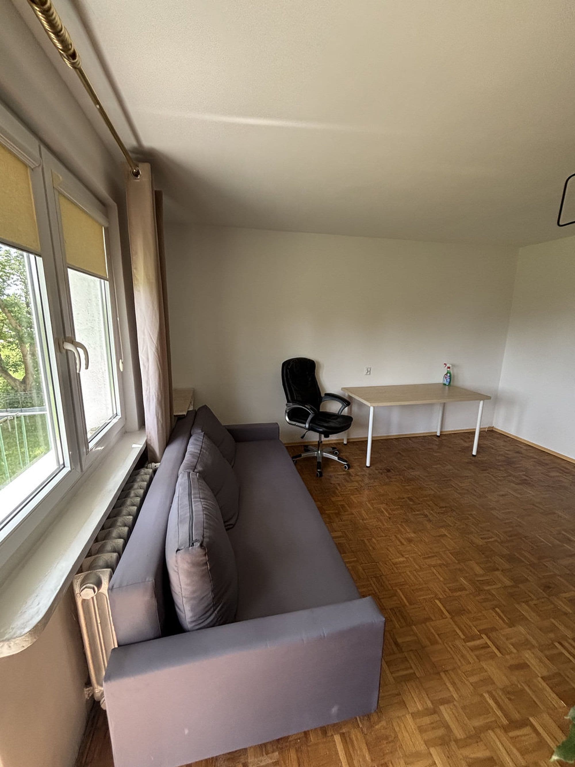 A room with a grey sofa, a black office chair, a wooden floor, and a window with curtains.