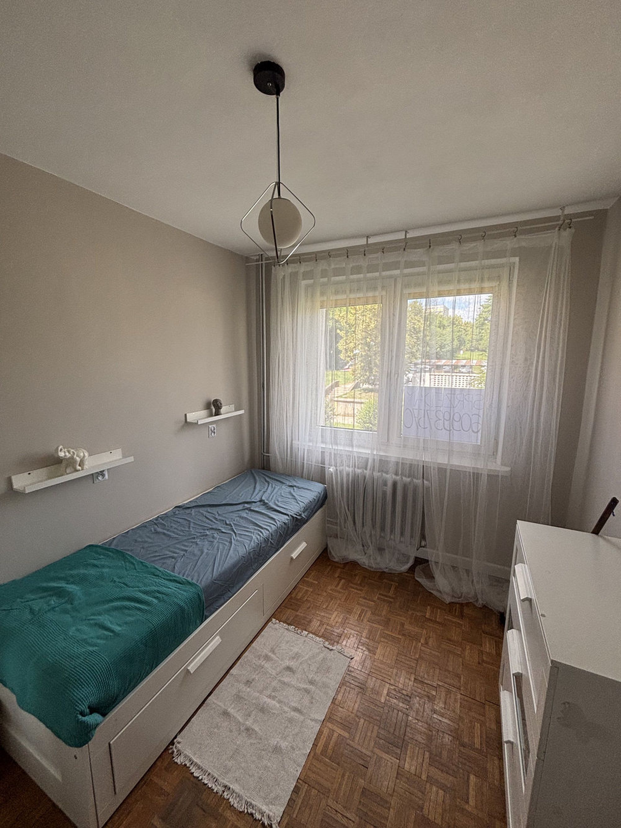 Une petite chambre avec un lit simple, un plancher en bois, une fenêtre avec des rideaux légers, une table de chevet et un luminaire au plafond.