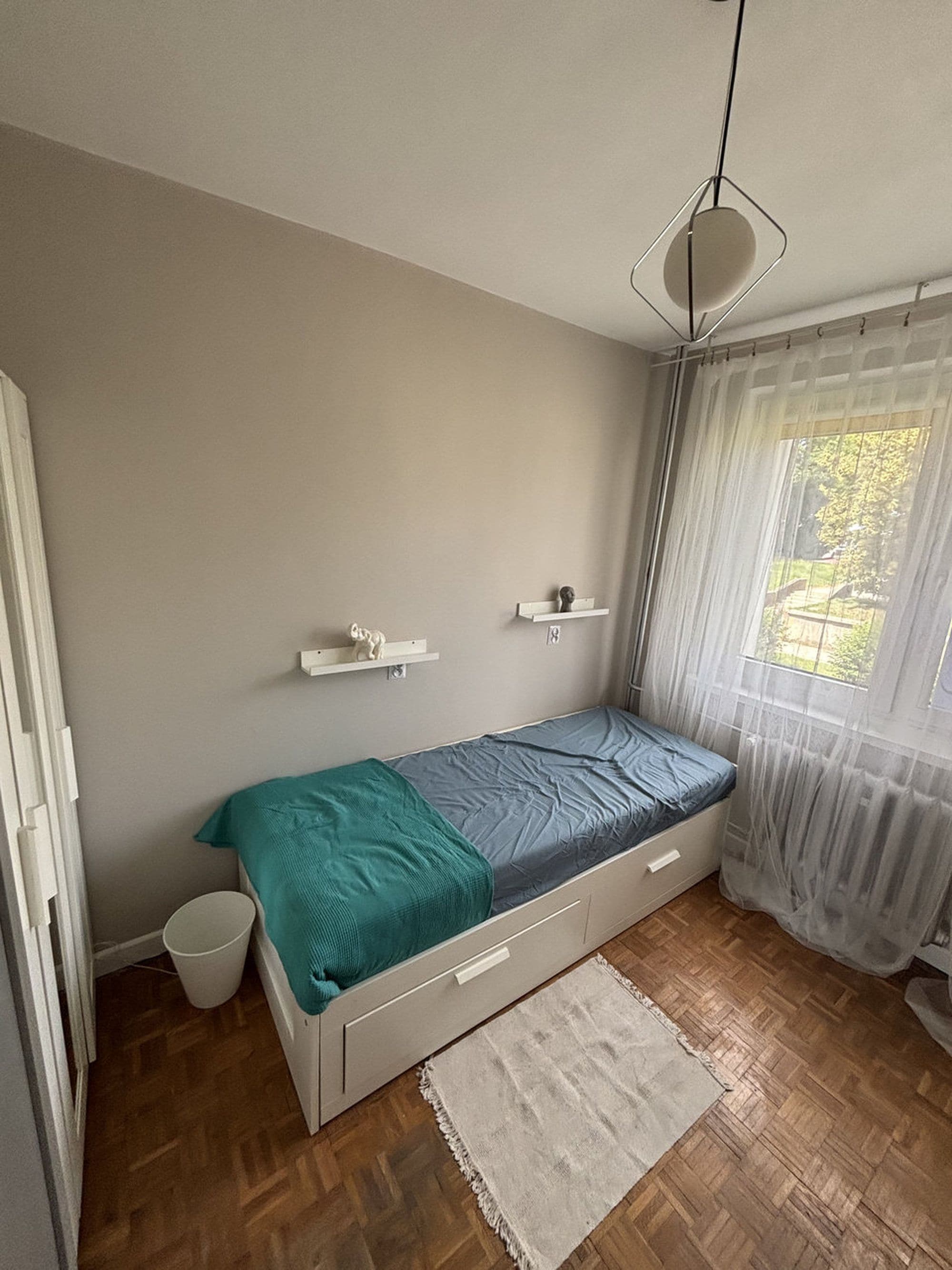Een slaapkamer met een bed, een klein wit kastje, een witte prullenbak en een raam met dunne gordijnen.