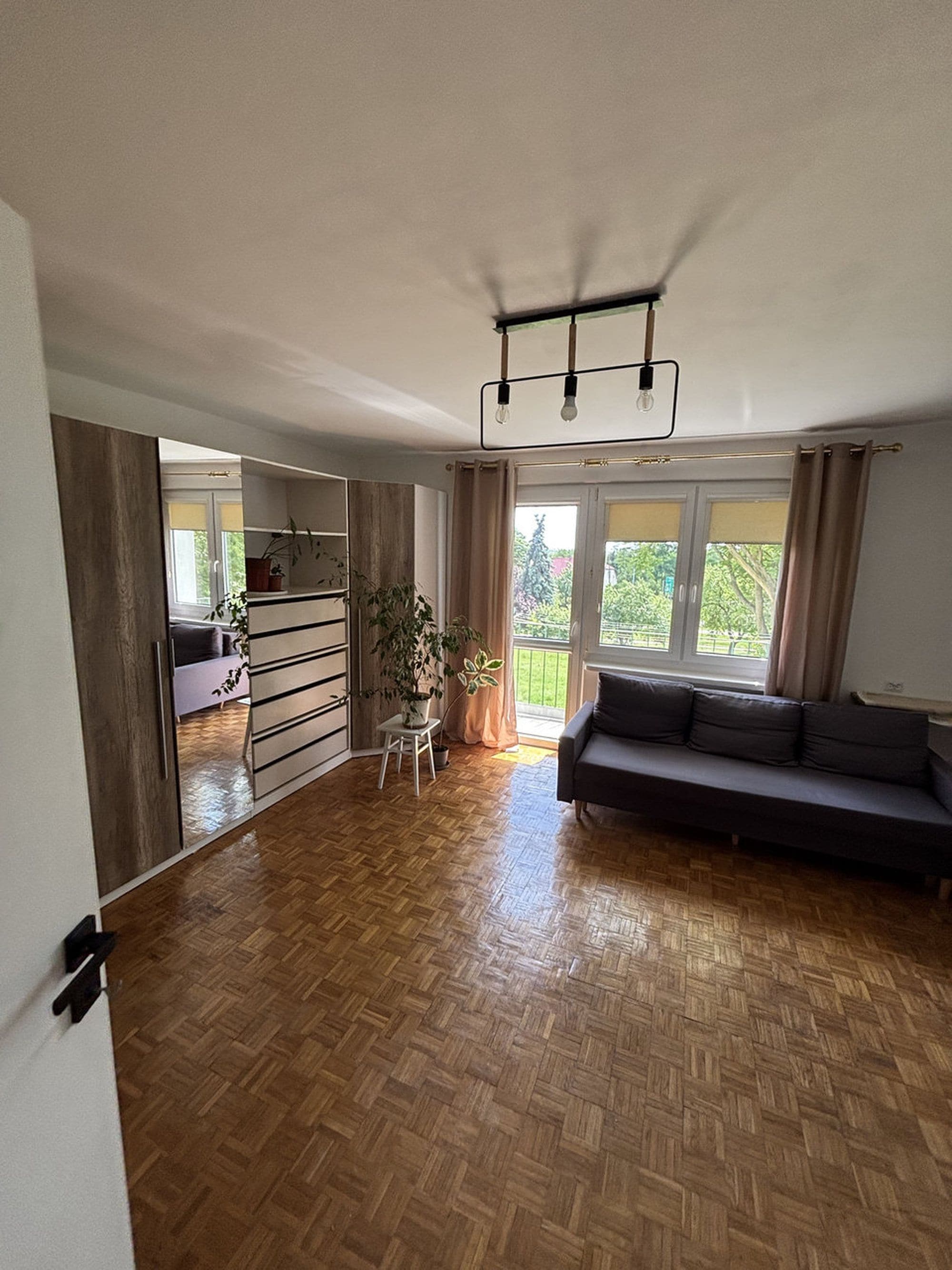A spacious living area with a wooden parquet floor, a dark sofa, a small table, and a potted plant.