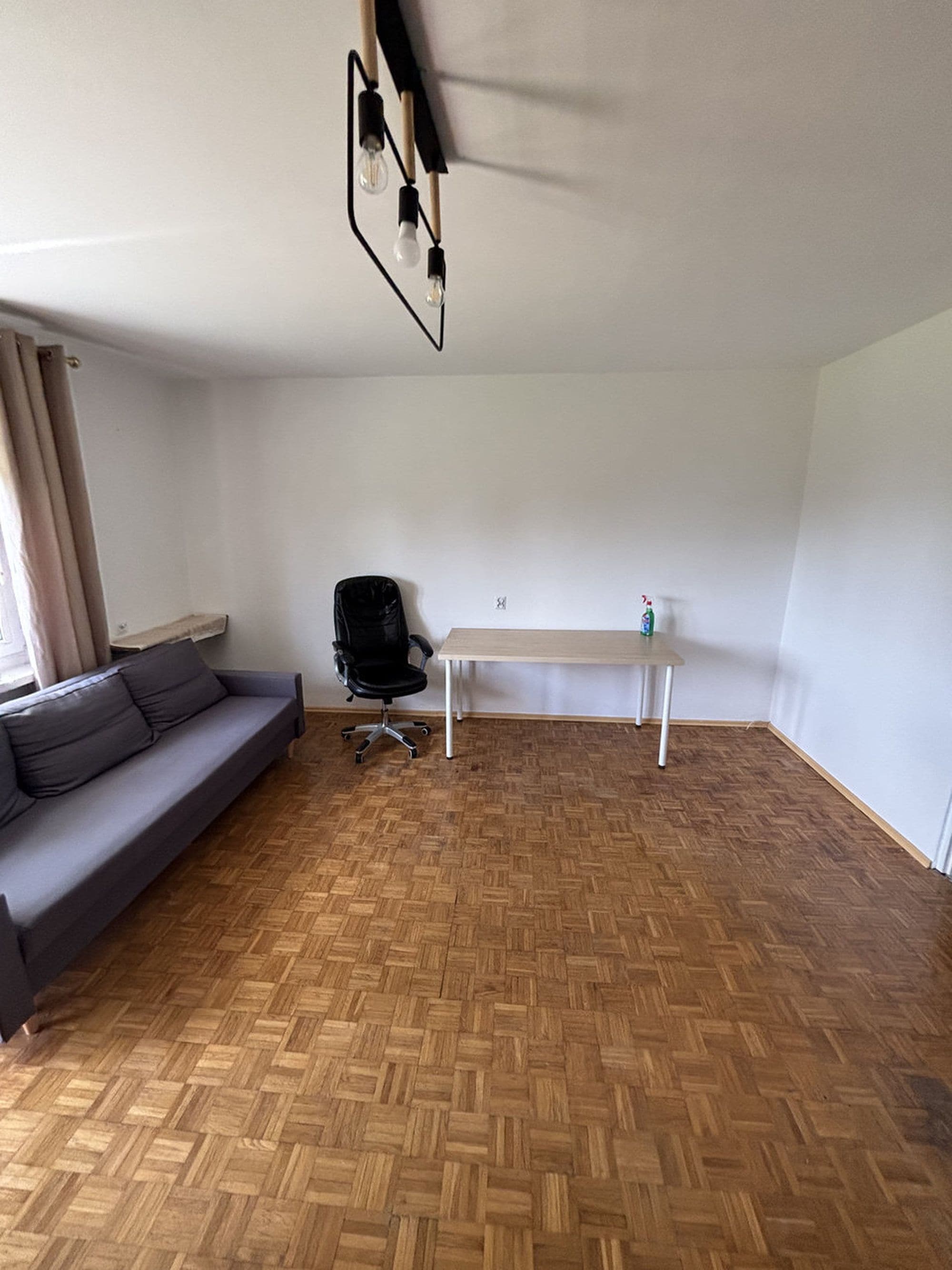 A room with a herringbone wooden floor, a grey sofa, a black office chair, and a white table.
