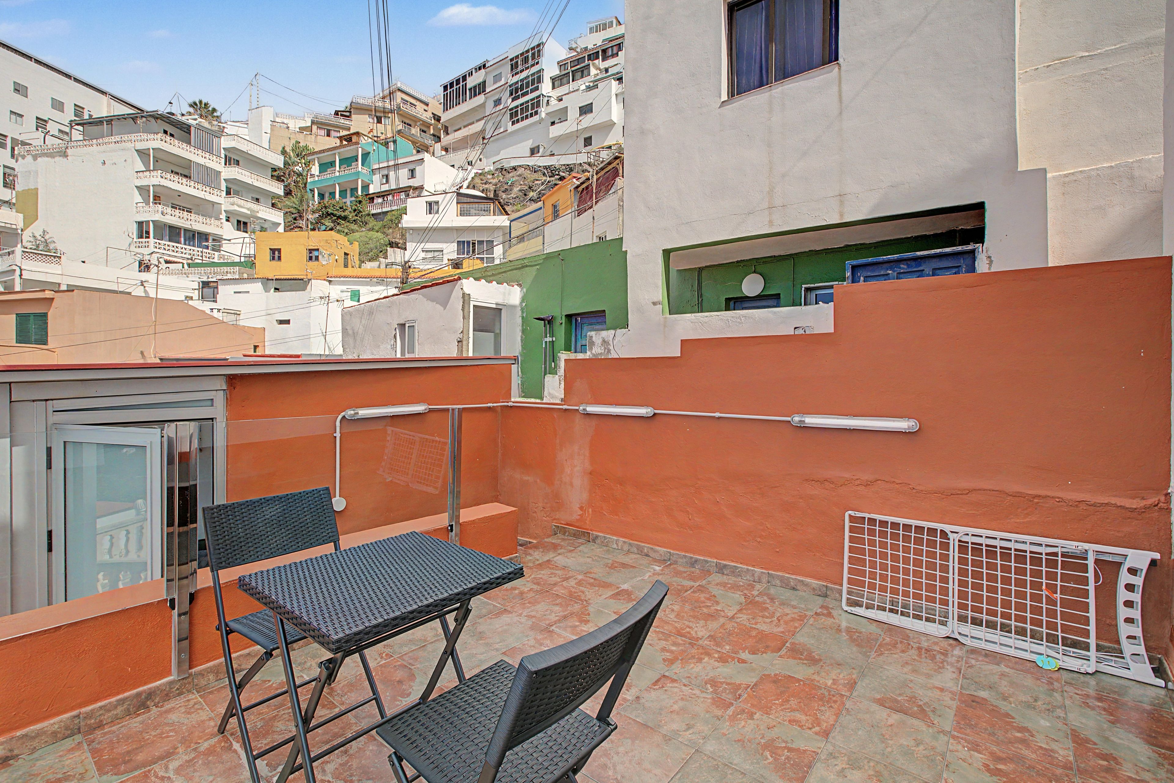 A rooftop terrace with a table and chairs, a metal railing, and a view of surrounding buildings.