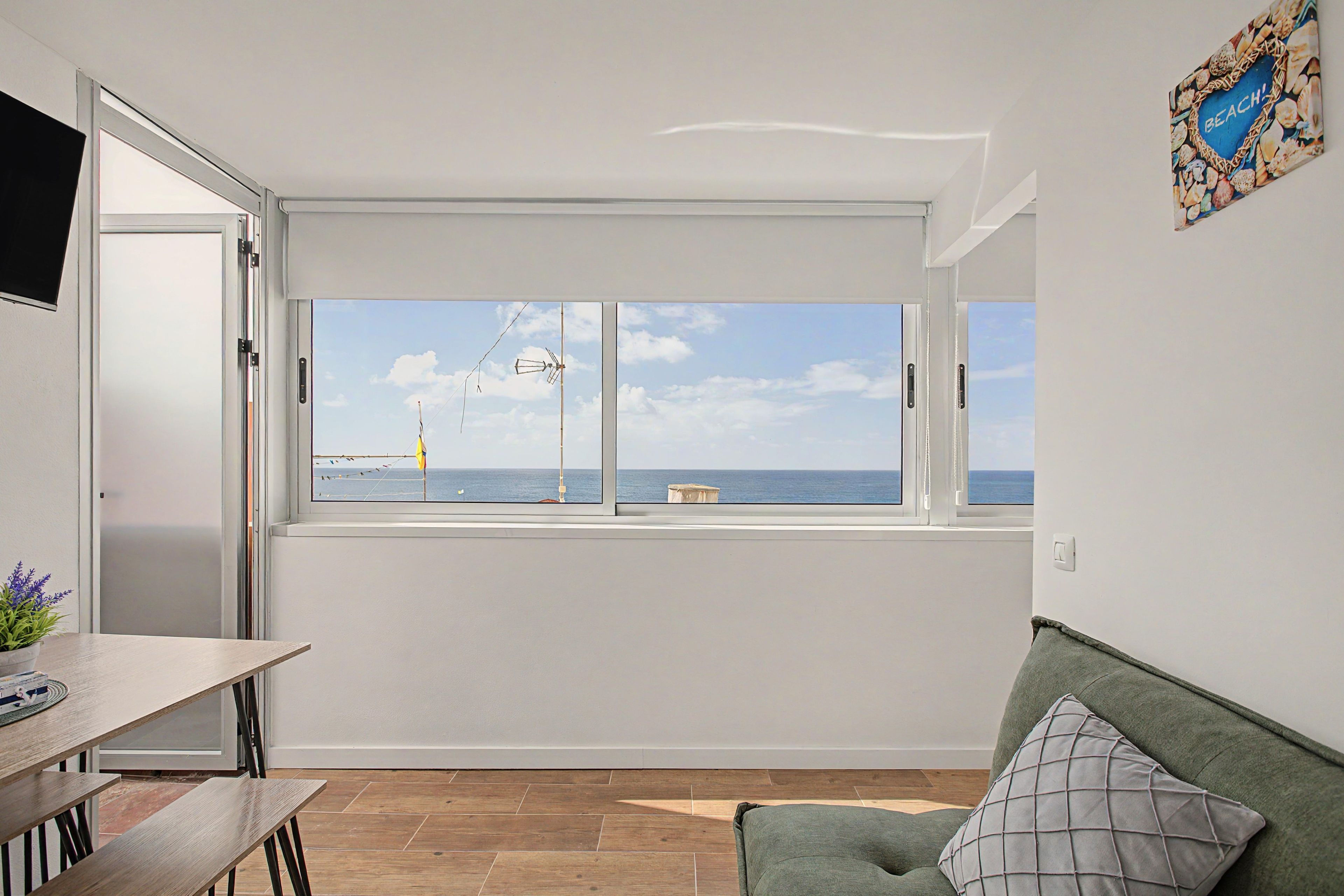 A room with a large window overlooking the sea, a wooden table, a green sofa, and a wall-mounted TV.
