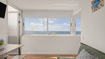 A room with a large window overlooking the sea, a wooden table, a green sofa, and a wall-mounted TV.