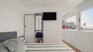 Ein Schlafzimmer mit einem Bett, einem an der Wand montierten Fernseher, einer kleinen Ablagefläche mit Gegenständen und einem Fenster mit Blick auf Gebäude.