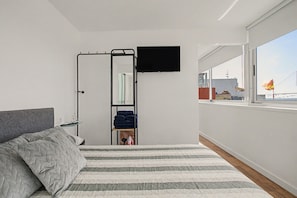 Een slaapkamer met een bed, een aan de muur bevestigde televisie, een klein plankje met spullen en een raam met uitzicht op gebouwen.