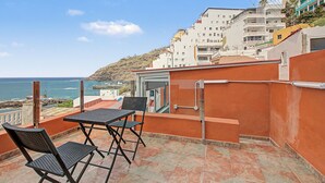 Ein Balkon mit Tisch und Stühlen mit Blick auf die Küste, im Hintergrund sind Gebäude zu sehen.