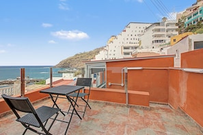 Een balkon met een tafel en stoelen, met uitzicht op de kust en gebouwen op de achtergrond.