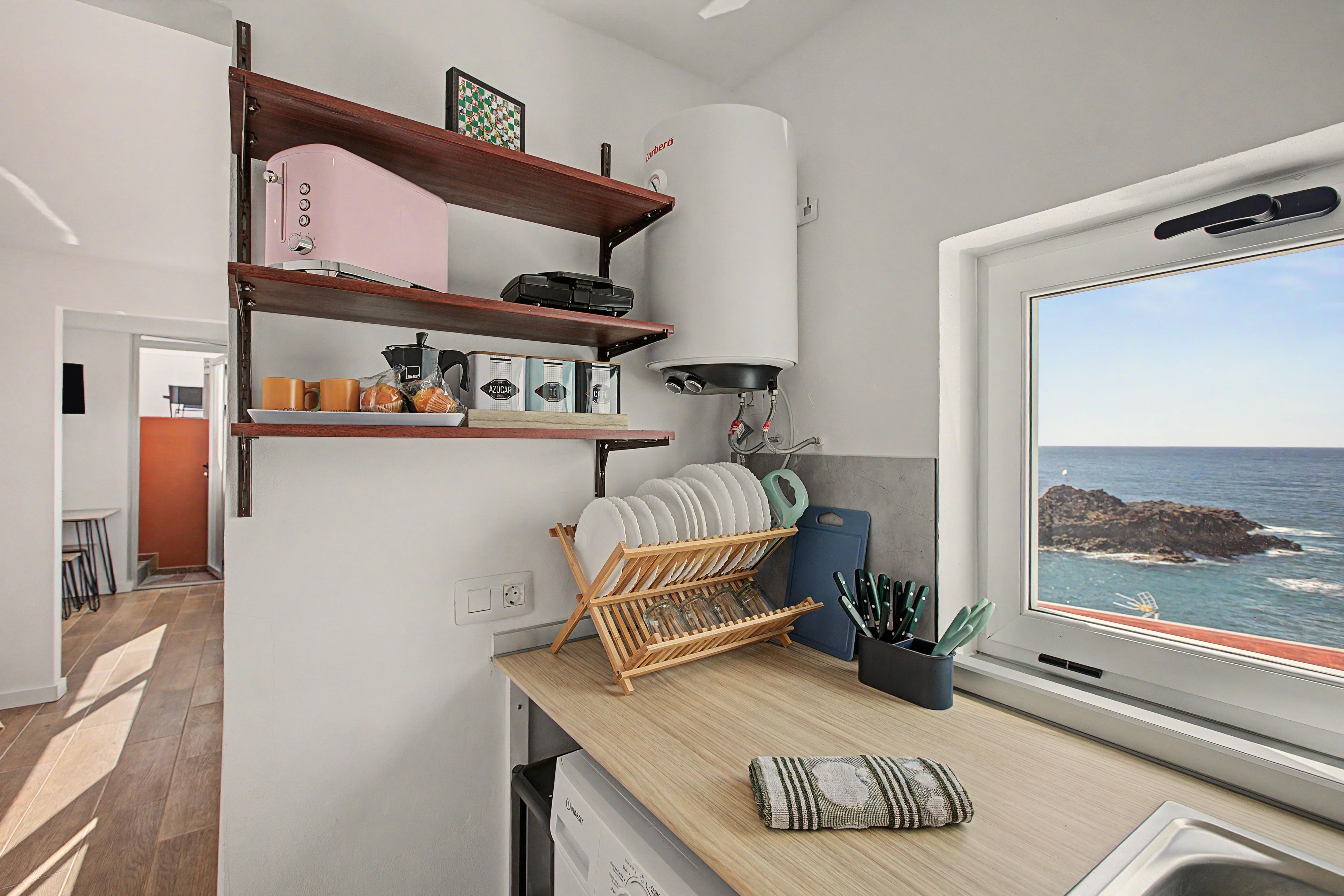 A kitchen with wooden shelves, a coffee machine, and a view of the sea through a window.