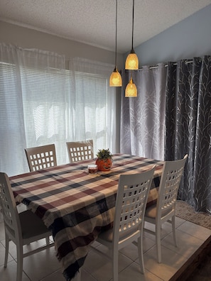 Un comedor con mantel a cuadros, sillas blancas, una planta en maceta y lámparas colgantes.
