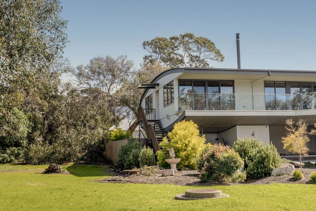 A modern two-story house with a balcony, surrounded by a well-maintained garden with various shrubs and a small fountain.