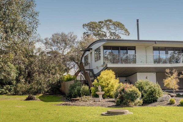 A modern two-story house with a balcony, surrounded by a well-maintained garden with various shrubs and a small fountain.