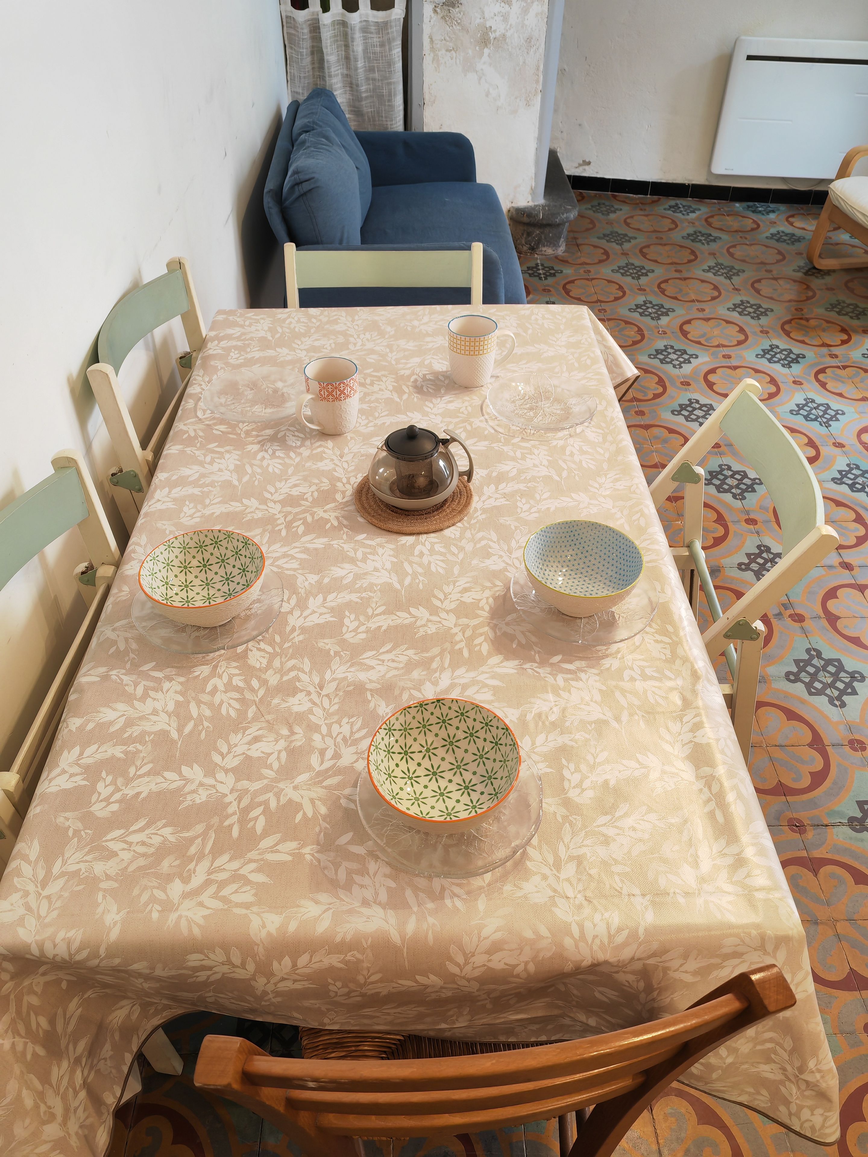 A dining table with a floral tablecloth, surrounded by wooden chairs, with a coffee pot and bowls on the table.