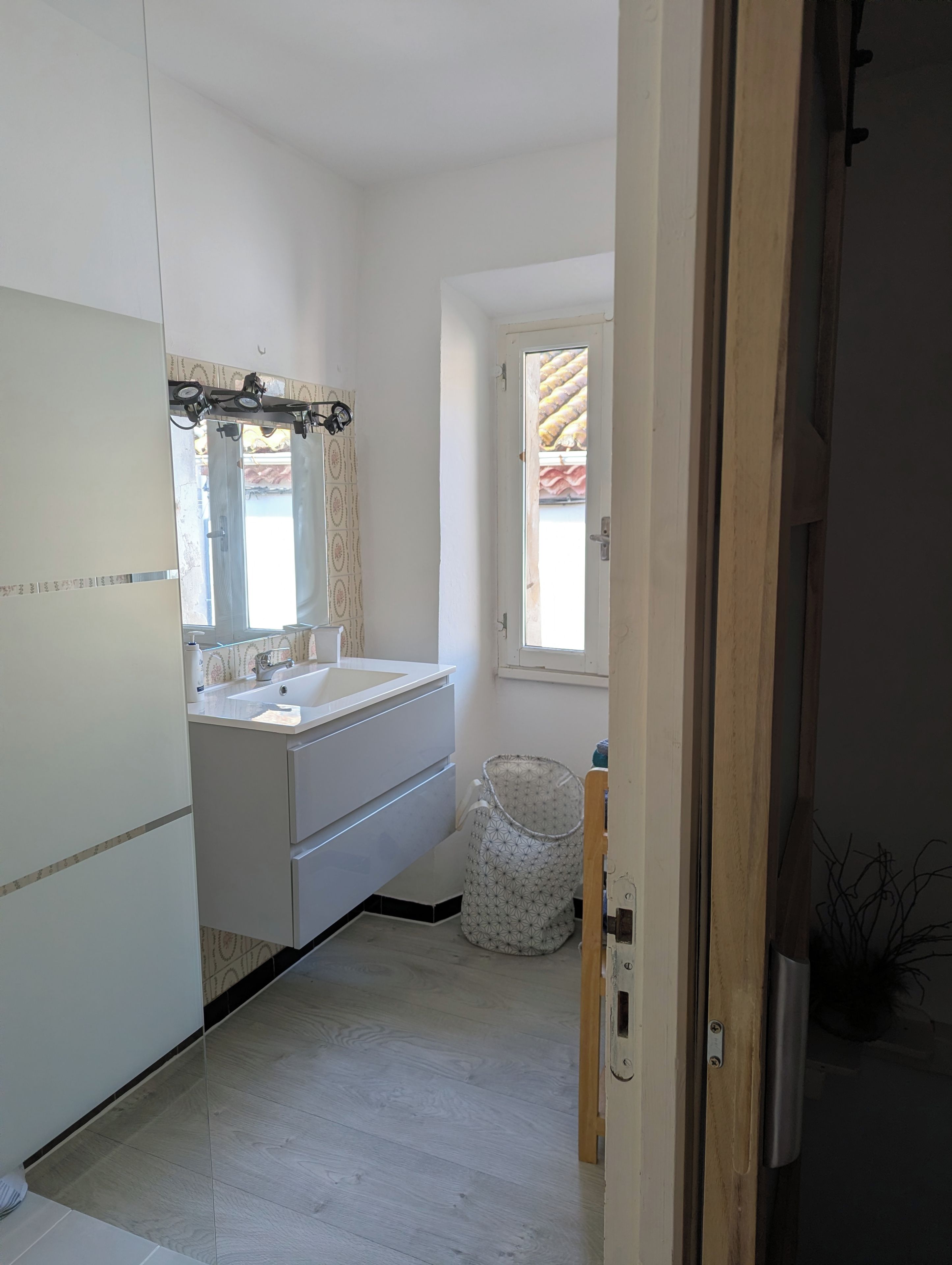 Une salle de bain avec un lavabo blanc, un miroir et des armoires. Il y a une fenêtre avec des rideaux et une vue sur un bâtiment à l’extérieur.
