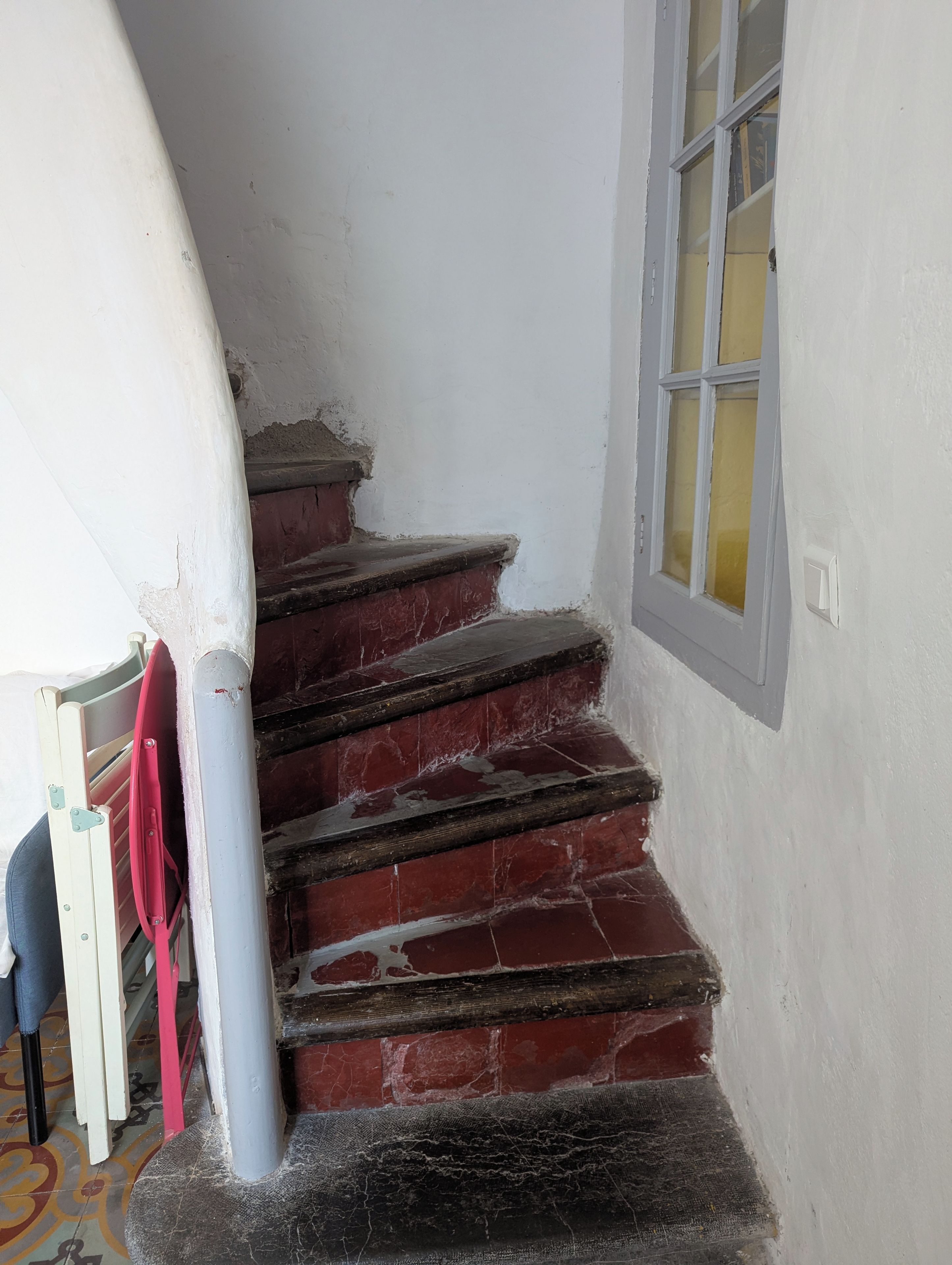 Un escalier aux marches rouges menant à un palier, un mur blanc et une fenêtre ornée d’un rideau jaune.