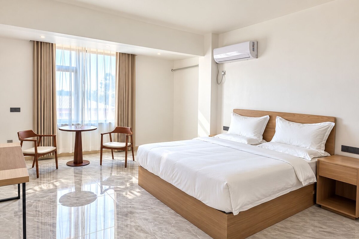 A hotel room with a bed, a bedside table, a chair, a small table, and a window with curtains.