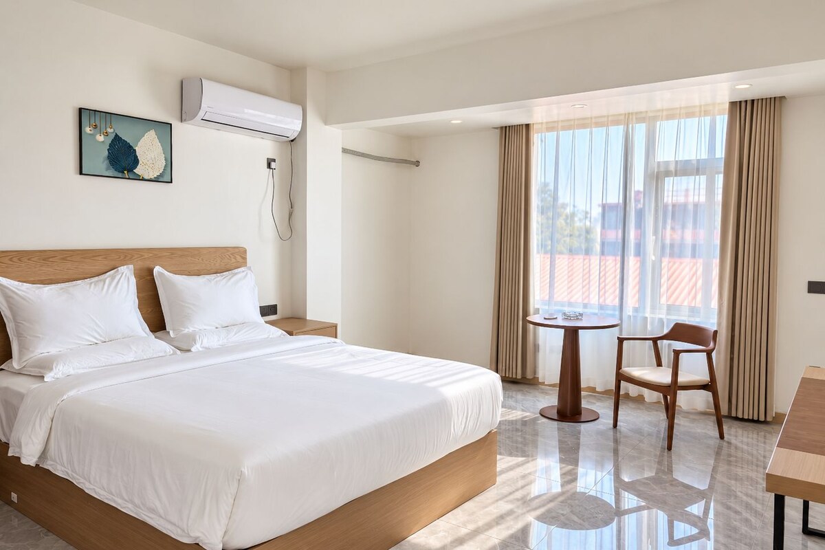 A hotel room with a bed, a bedside table, a chair, a small table, and a window with curtains.