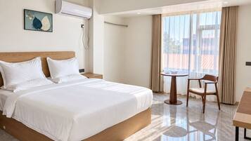 Una habitación de hotel con una cama, una mesita de noche, una silla, una pequeña mesa y un ventanal con cortinas.