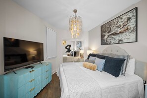 A bedroom with a large bed, a television, a blue dresser, a chandelier, and a painting on the wall. - Caribe BldgB-Fully Updated-Great Gulf Views-Orange Beach-Signature Properties (Orange Beach)