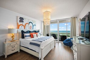 Een slaapkamer met een bed, nachtkastje, lamp, kunstwerk en uitzicht op de oceaan door grote ramen.