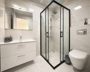 A modern bathroom with a glass-enclosed shower, a white toilet, a vanity with a mirror, and a light switch on the wall. - LUNA MARE REZİDANS (FİNİKE)