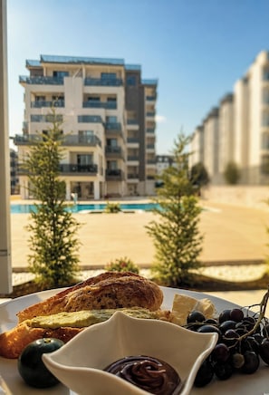 A meal with toast, avocado, and chocolate spread on a plate, with a pool and buildings in the background. - LUNA MARE REZİDANS (FİNİKE)