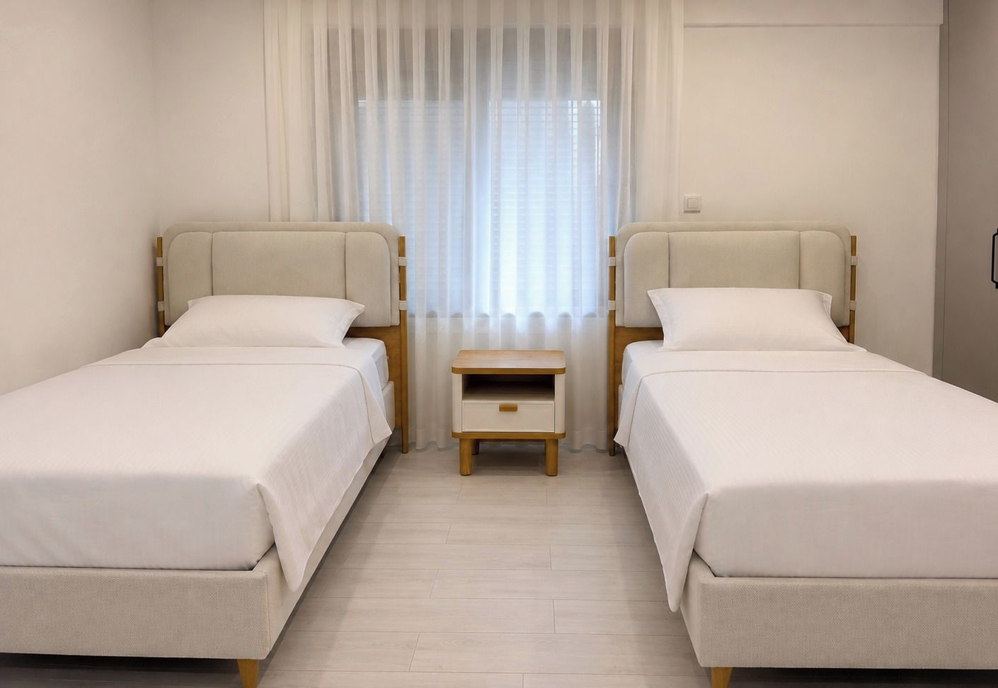 Una camera d'albergo con due letti, un comodino e una finestra con tende leggere.