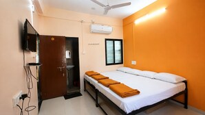 A hotel room with a bed, a TV mounted on the wall, an air conditioning unit, and a window with blinds. - Shri Radha Govind Bhakt Niwas and Hotel (Pandharpur)