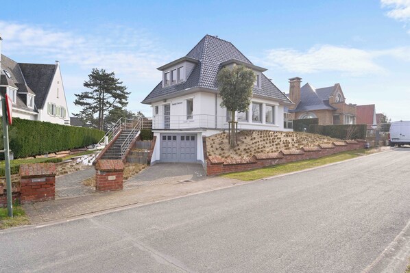 Ein zweistöckiges Haus mit einem steilen Satteldach, einem Hauseingang mit Doppeltüren und einem kleinen Vorgarten mit Baum und Ziegelmauer.