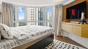 Une chambre moderne avec un grand lit, un téléviseur fixé au mur et une vue sur de hauts immeubles, visible à travers de grandes fenêtres.