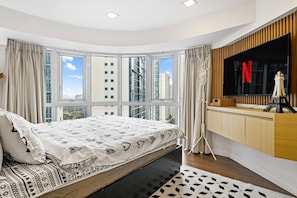 Ein modernes Schlafzimmer mit einem großen Bett, einem an der Wand montierten Fernseher und Blick auf hohe Gebäude durch große Fenster.