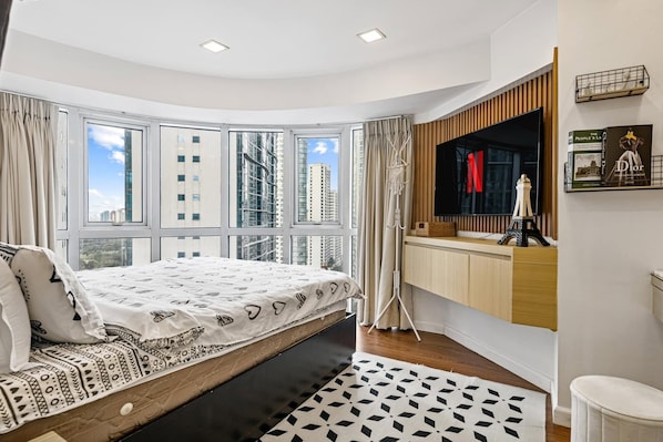 Ein modernes Schlafzimmer mit einem Bett, einem großen Fenster, einem Schminktisch mit Spiegel und Blick auf hohe Gebäude.