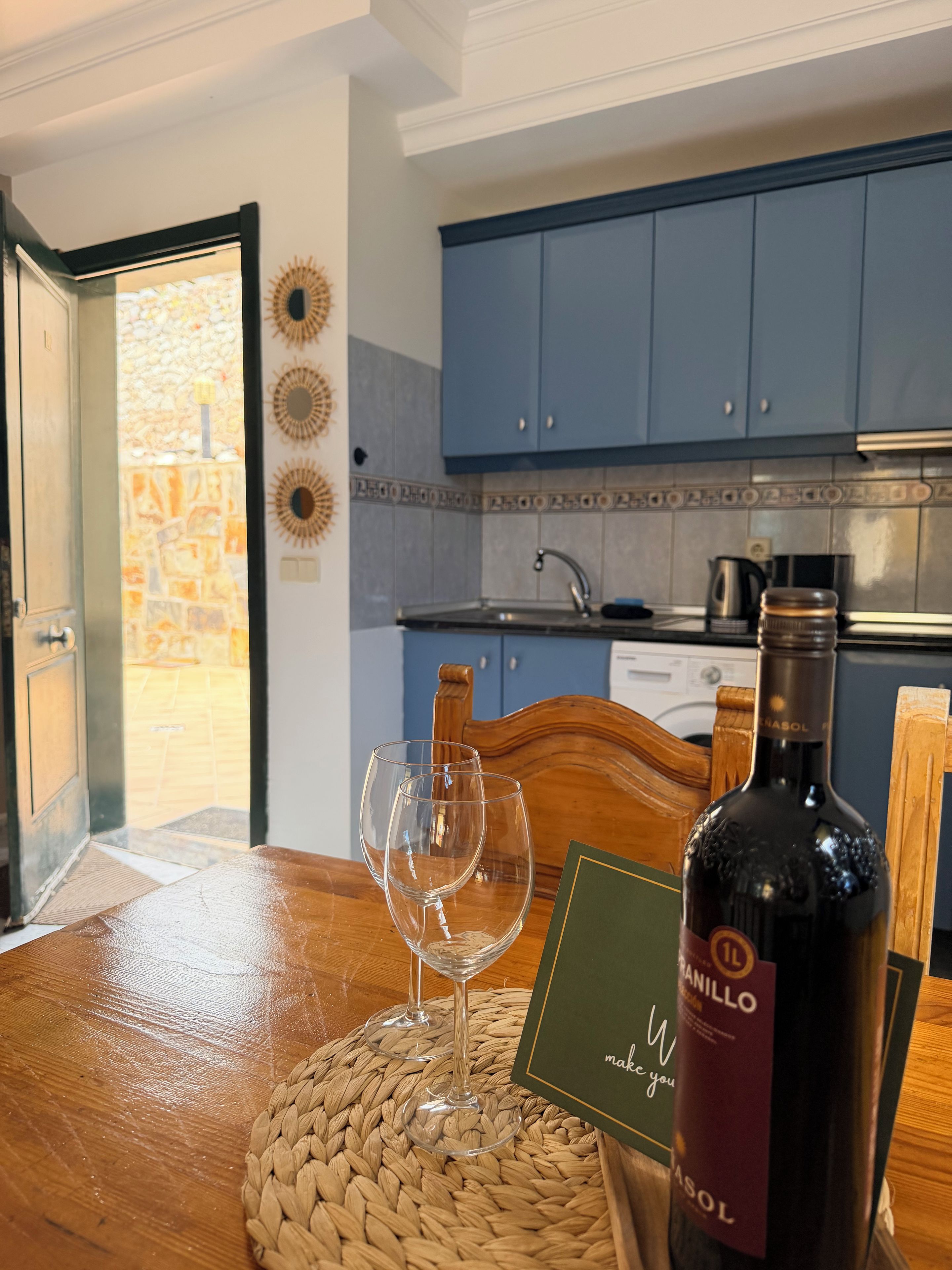 Een keuken met blauwe kastjes, een houten tafel met een geweven placemat, twee wijnglazen en een fles wijn.