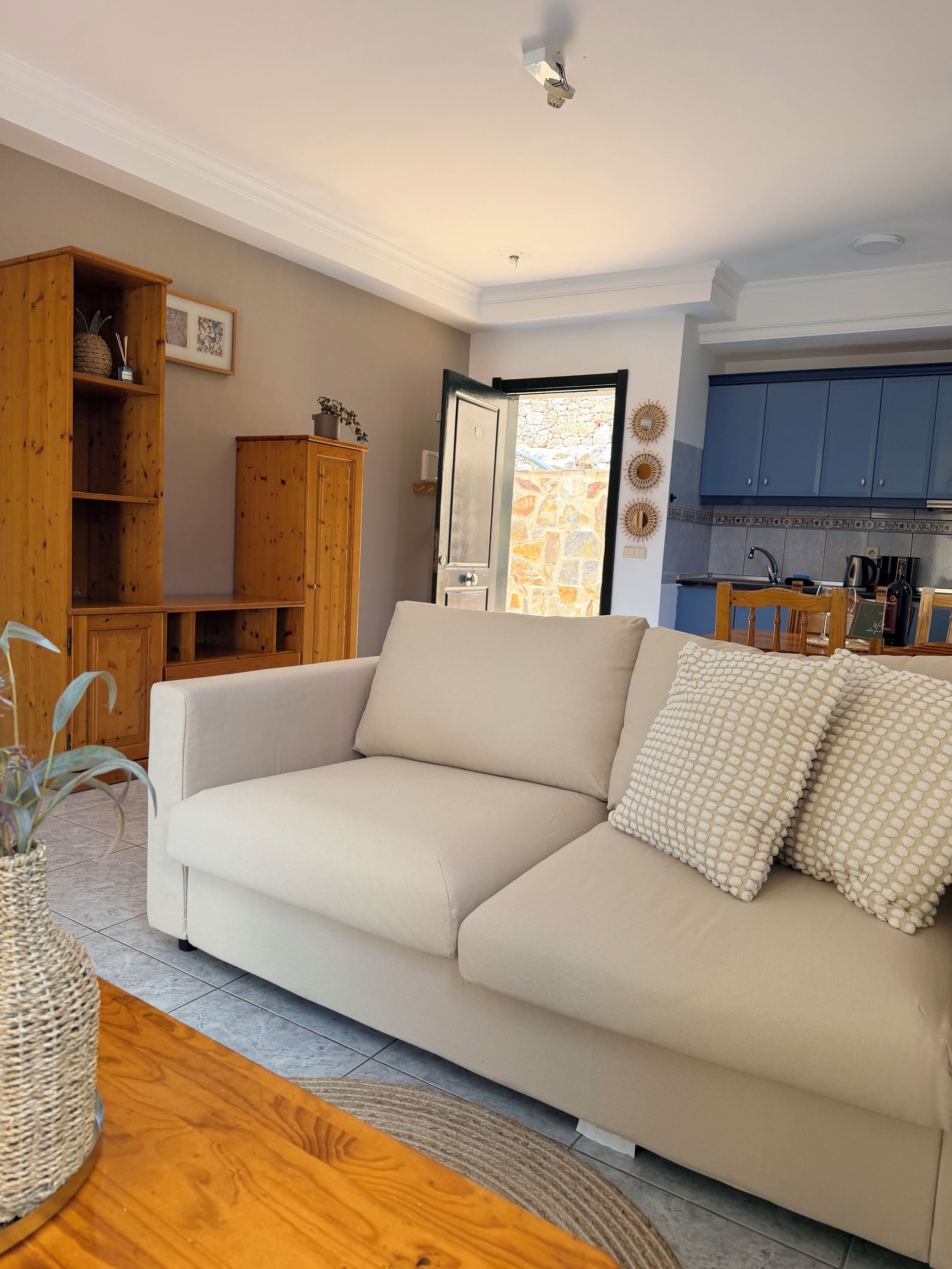 Ein Wohnzimmer mit einem beigen Sofa, einem Holzschrank und einem durch eine offene Tür einsehbarer Küchenbereich.